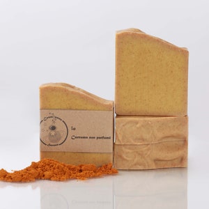 Peut inclure: Quatre savons jaunes et orange empilés les uns sur les autres. Le savon du haut porte une étiquette en papier brun avec le texte "lo Curcuma non parfumé" et une petite illustration d'une abeille en combinaison spatiale. Un tas de poudre de curcuma orange se trouve devant le savon.