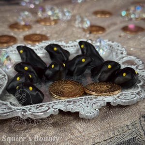 May include: A decorative silver tray holding several black, stylized creature figures with yellow eyes. Two gold-coloured, circular coins are also present. The background includes scattered crystals and coins, with the text "Squire's Bounty" at the bottom.