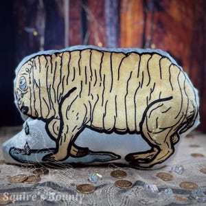 May include: A plush pillow shaped like a light-yellow creature with black outlines. The pillow has a light blue base with a drawing of a skull and water. The words "Squire's Bounty" are at the bottom.