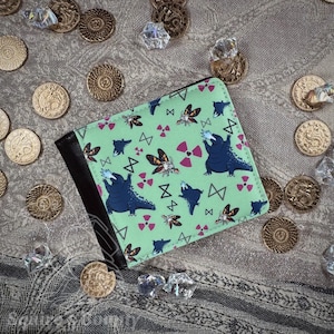 May include: A bi-fold wallet with a light green background and a pattern of dark blue Godzilla-like creatures, moths, and pink radiation symbols. The wallet has a black leather interior and a money clip.