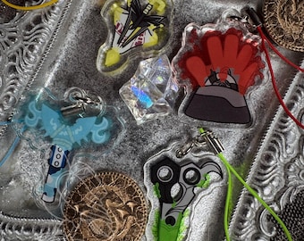 May include: A selection of acrylic charms and a faceted crystal. The charms display diverse designs, such as a sword, scissors, and a stylised volcano. The charms have coloured cords attached for hanging. Two gold-coloured coins are also visible.