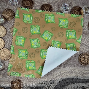 May include: A brown fabric featuring a repeating pattern of green, blue, and yellow square designs and geometric shapes. The fabric is partially folded, showing a white underside. The image is surrounded by coins and crystals.