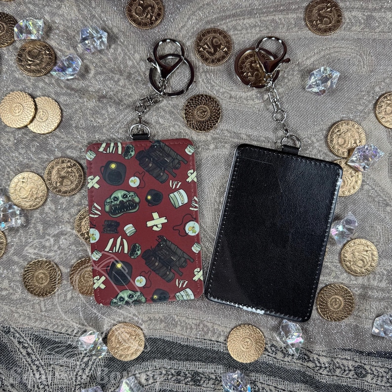 May include: Two keychains with cardholders. One cardholder is red with a repeating pattern of skulls, weapons, and other symbols. The other cardholder is black. Both have silver-colored key rings and chains.