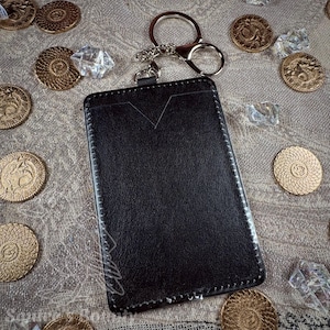 May include: A black leather cardholder with a silver keychain and clasp. The cardholder has a V-shaped cutout at the top and white stitching around the edges. Gold coins and clear crystals are scattered around the cardholder.