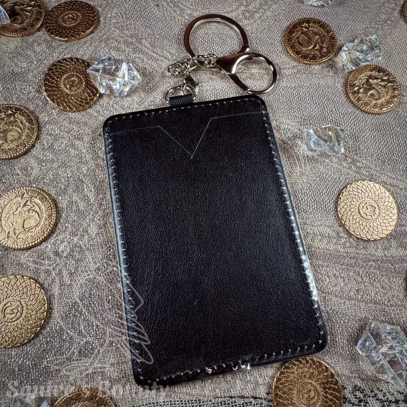 May include: A black leather cardholder with a silver keychain and clasp. The cardholder has a V-shaped cutout at the top and white stitching around the edges. Gold coins and clear crystals are scattered around the cardholder.