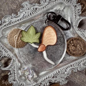 May include: A clear acrylic keyring with a light green leaf and a tan seed design. The keyring has a black clasp and is displayed on a decorative silver tray with gold coins.