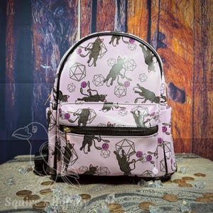 May include: A light pink backpack featuring a repeating pattern of black cats playing with purple yarn balls and geometric polyhedral shapes. The bag includes a front zippered pocket, black trim, and gold-toned hardware on the zipper pull.