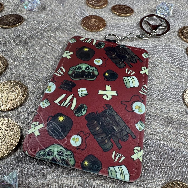 May include: A burgundy cardholder with a repeating pattern of monster faces, barrels, and other graphics. The cardholder has a silver key ring attached. Gold coins and crystals are scattered around the cardholder.