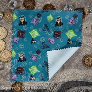 May include: A teal fabric square featuring a repeating pattern of cartoon owls, octopuses, and fantasy creatures. The fabric has a white underside and is surrounded by gold coins and crystals.