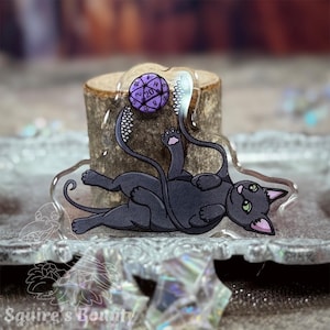 May include: A black cat with green eyes is playing with a purple 20-sided die. The cat is lying on its back with its paws in the air. The die is attached to a string that is wrapped around the cat's tail.