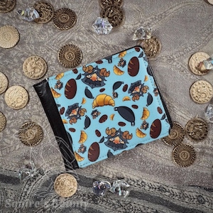 May include: A bi-fold wallet with a light blue background featuring a pattern of cartoon characters, croissants, and bread. The wallet has a black leather exterior and is surrounded by gold coins and crystals.