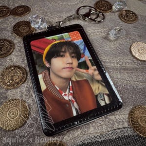May include: Black ID card holder with a photo of a person, a silver keychain, and a collection of gold coins and clear crystals. The person in the photo is wearing a yellow beanie and a brown jacket.