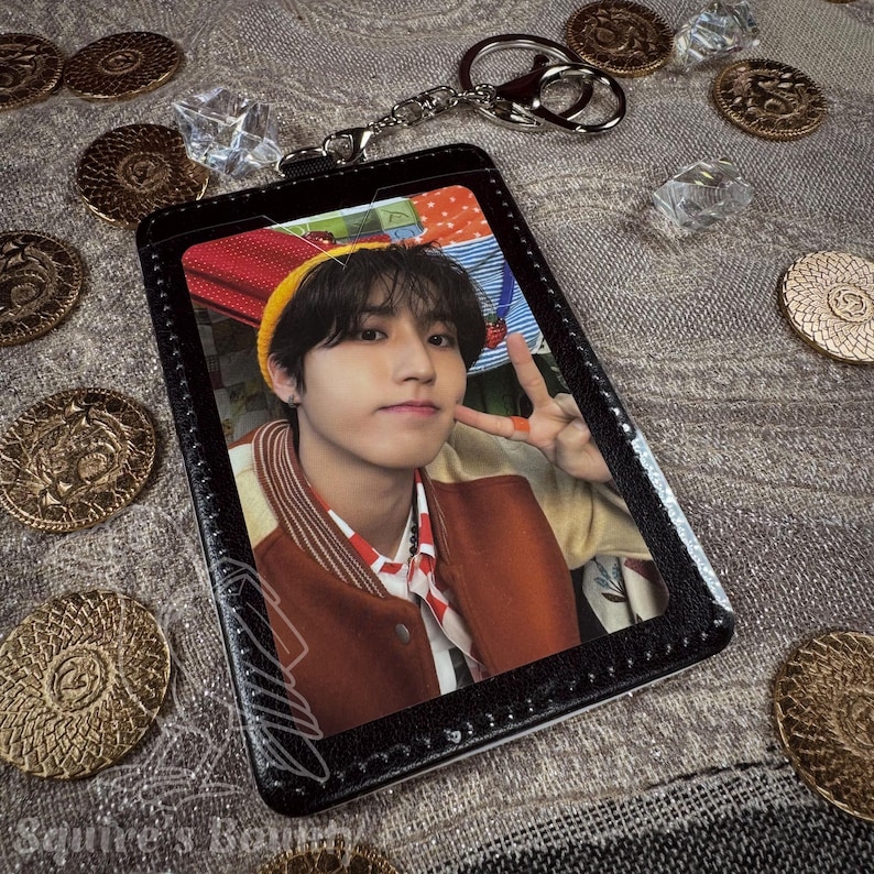 May include: Black ID card holder with a photo of a person, a silver keychain, and a collection of gold coins and clear crystals. The person in the photo is wearing a yellow beanie and a brown jacket.