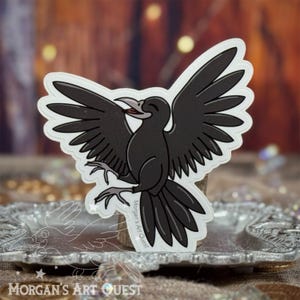May include: A sticker featuring a cartoon crow with outstretched wings. The crow is black with a white outline, and its beak is open. The sticker has the text "Morgan's Art Quest".