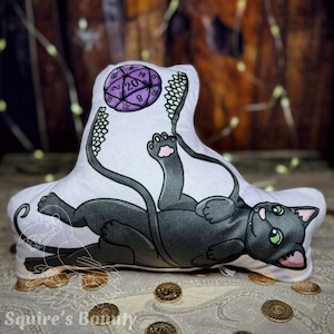 May include: A black cat plush toy with a purple 20-sided die in its paw. The cat is lying on its back with its legs in the air. The plush toy is white with black details and has a green eye. The die is a standard D20 with numbers 1-20.