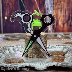 May include: A clear acrylic scissor-shaped keychain with black and white blades and a green handle. The keychain is attached to a green clasp and is displayed on a small wooden stand. The text "Squire's Bounty" is visible at the bottom.