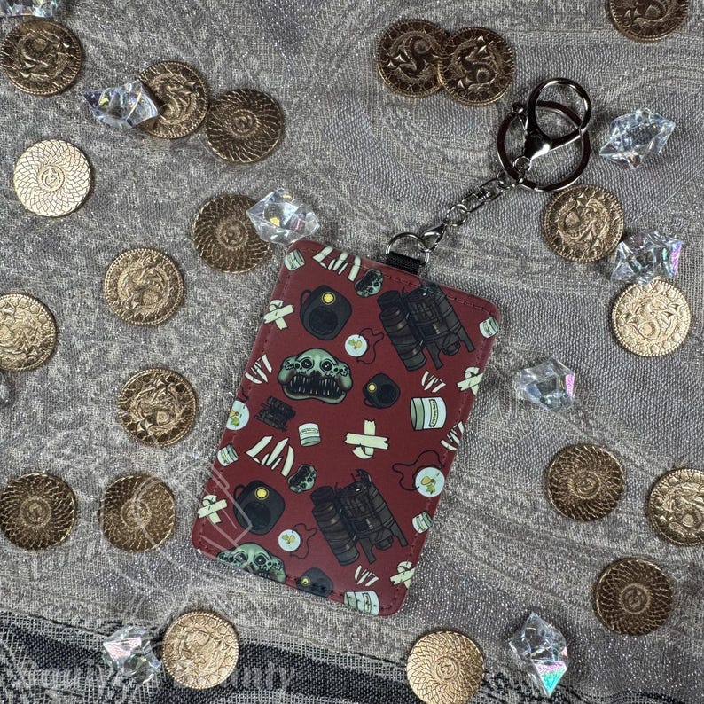 May include: A burgundy cardholder with a keychain, featuring a repeating pattern of skulls, teapots, and other graphics. The cardholder is surrounded by gold coins and clear crystals on a textured gray surface.