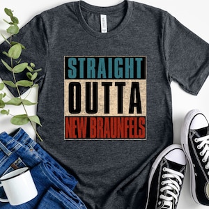 May include: A dark heather grey t-shirt with the text "STRAIGHT OUTTA NEW BRAUNFELS" in a vintage style font. The text is in a red, white, and blue color scheme.
