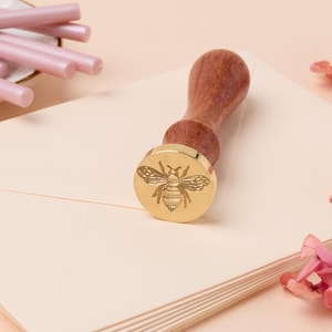 May include: A gold-toned wax seal stamp with a bee design. The stamp is attached to a wooden handle.