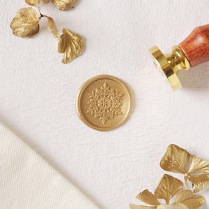 May include: A gold wax seal with a snowflake design. The seal is on a white surface with gold leaf accents.