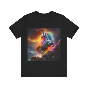 Könnte beinhalten: Ein schwarzes T-Shirt mit einem Motiv einer weißen Einhorn mit einer Regenbogenmähne und einem Regenbogen-Schwanz, das über eine felsige Bergkette springt.