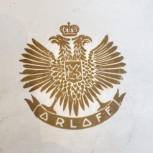 May include: A gold-colored crest with two eagles facing each other, a crown on top, and the word "ORLOFF" written below.