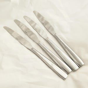 Puede incluir: Un juego de cinco cuchillos de mesa de acero inoxidable. Cada cuchillo tiene una hoja larga y delgada y un mango redondeado. Los cuchillos están dispuestos en diagonal sobre una superficie blanca.