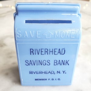 May include: A light blue plastic piggy bank with a slot on the top. It says "SAVE MONEY" on the front, "Riverhead Savings Bank" and "Riverhead, N.Y." below that, and "Member F.D.I.C." at the bottom.