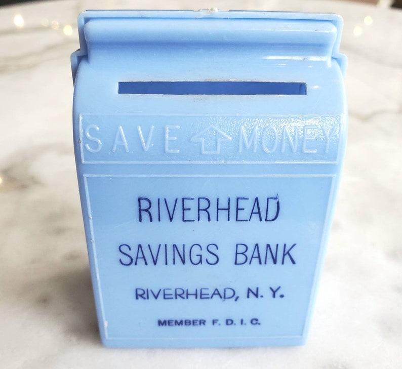 May include: A light blue plastic piggy bank with a slot on the top. It says "SAVE MONEY" on the front, "Riverhead Savings Bank" and "Riverhead, N.Y." below that, and "Member F.D.I.C." at the bottom.