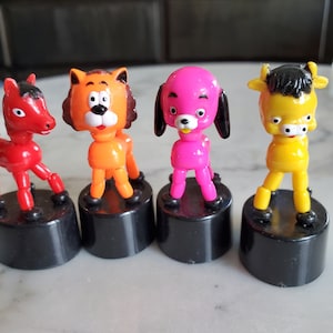 May include: Four colorful bobblehead figurines on black bases. The figurines are a red horse, an orange cat, a pink dog, and a yellow cow.