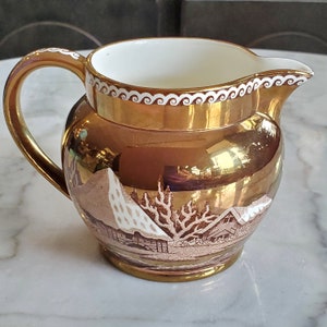 May include: A gold-toned ceramic creamer with a white and brown design of a winter scene. The creamer has a white rim with a decorative swirl pattern.