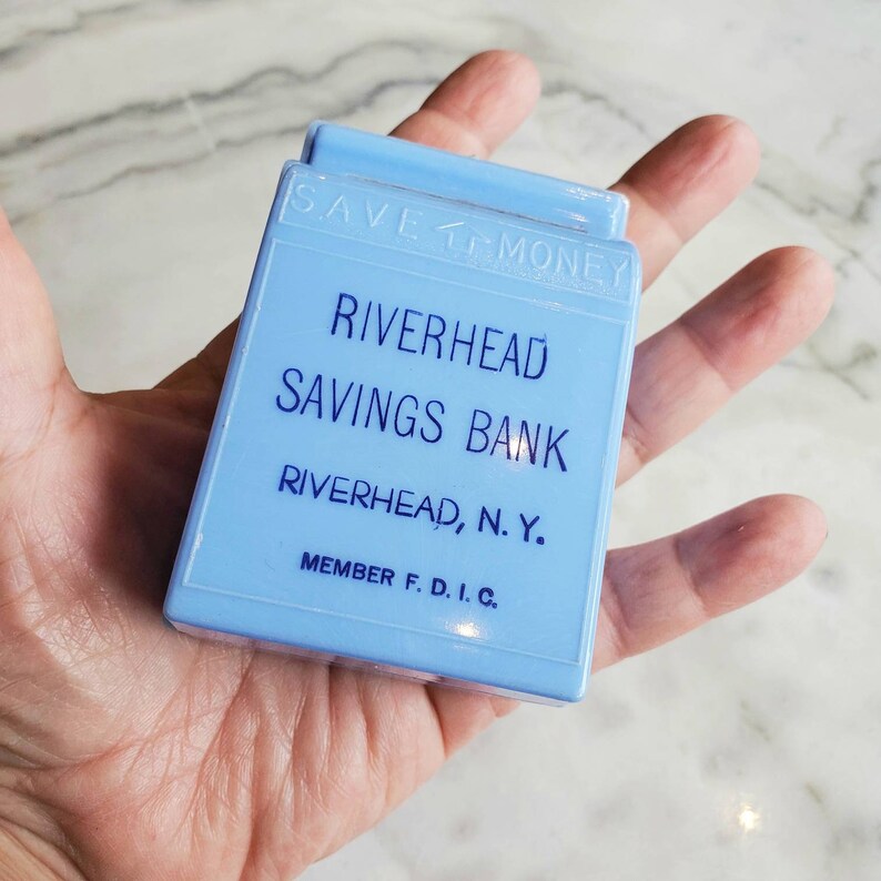May include: A light blue piggy bank with the text "SAVE MONEY" on the top, "RIVERHEAD SAVINGS BANK" in the center, and "RIVERHEAD, N.Y. MEMBER F.D.I.C." at the bottom.