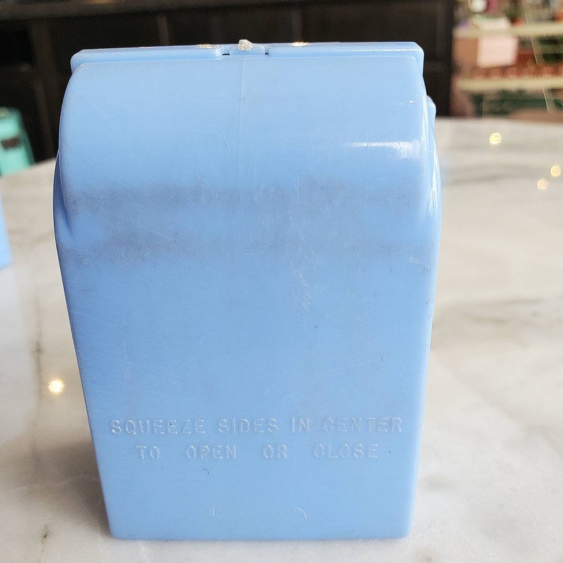 May include: A light blue plastic container with the text "Squeeze sides in center to open or close".