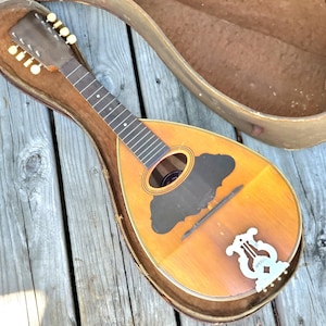 Peut inclure: Mandoline ancienne en bois clair, avec un trou sonore sombre. L'instrument est dans son étui ouvert et usé, posé sur une surface en bois vieilli. La mandoline présente une incrustation blanche décorative près du chevalet.