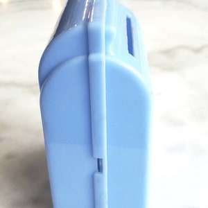 May include: A light blue plastic piggy bank with a slot on the top for coins. The bank is closed and has a seam down the side.