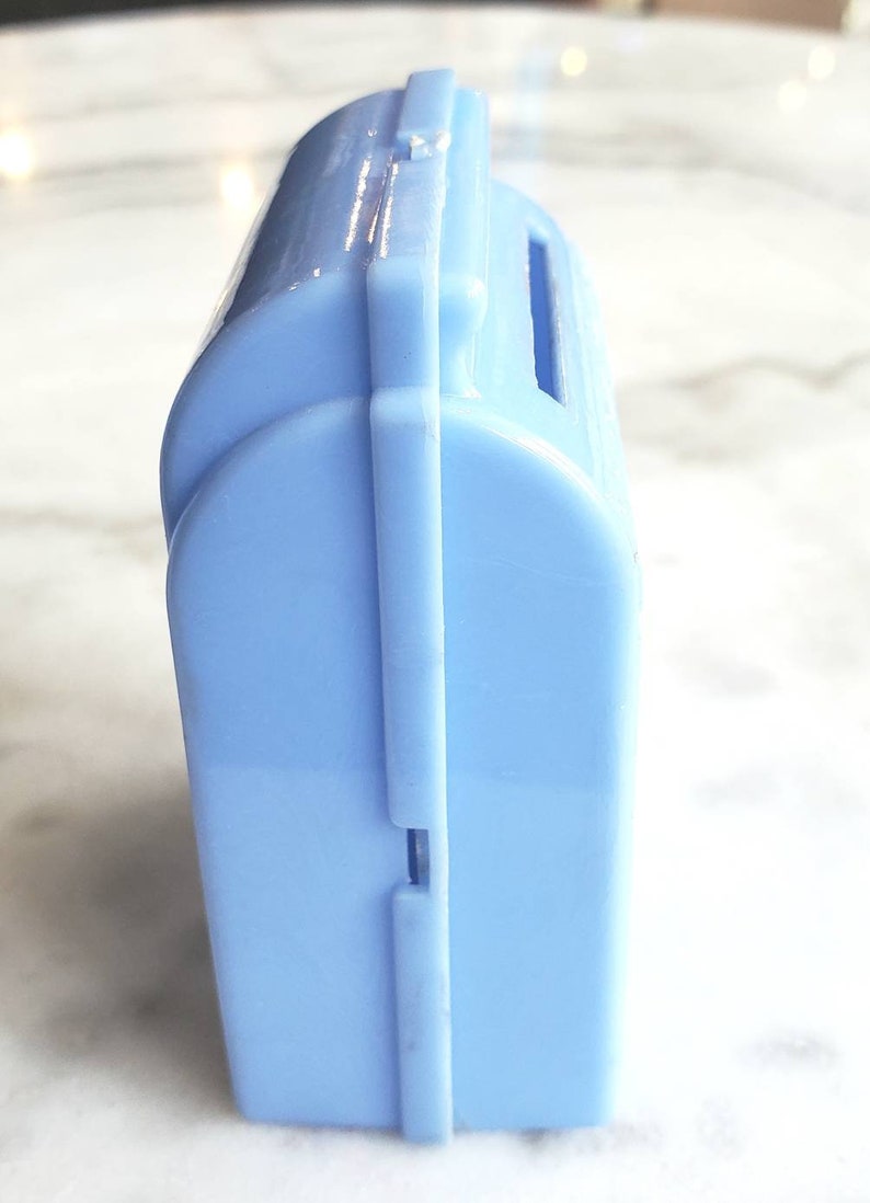 May include: A light blue plastic piggy bank with a slot on the top for coins. The bank is closed and has a seam down the side.