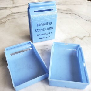 May include: A light blue plastic piggy bank with the text "Save Money" on the front. The bank is labeled "Riverhead Savings Bank, Riverhead, N.Y. Member F.D.I.C."