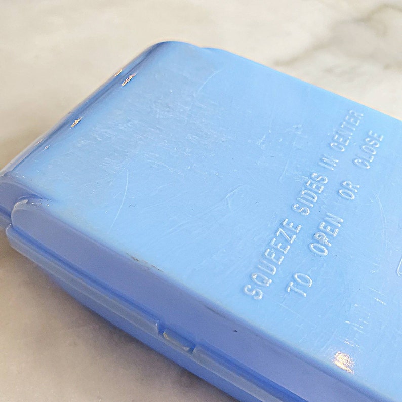 May include: A light blue plastic container with the text "Squeeze sides in center to open or close" embossed on the top.