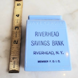 May include: A light blue ceramic piggy bank with the words "Save Money" on the top, "Riverhead Savings Bank" in the center, and "Riverhead, N.Y. Member F.D.I.C." at the bottom.