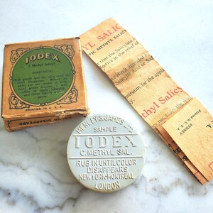 May include: A vintage Iodex Methyl Salicyl ointment set including a square tan cardboard box with a green circular label, a round light gray metal tin with embossed text, and a long, folded tan paper instruction leaflet with black text.