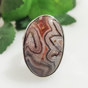 May include: Oval ring with a polished agate stone, displaying brown, white, and reddish-orange banding. The stone is set in a silver-coloured metal band. The ring's design highlights natural patterns and colours.