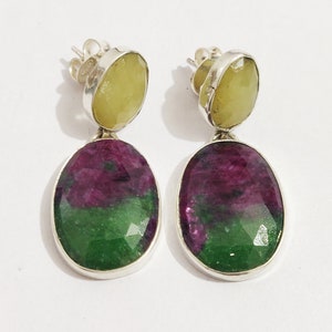 May include: A pair of silver earrings with green and purple oval-shaped gemstones. The top of each earring features a smaller, round, yellow gemstone.