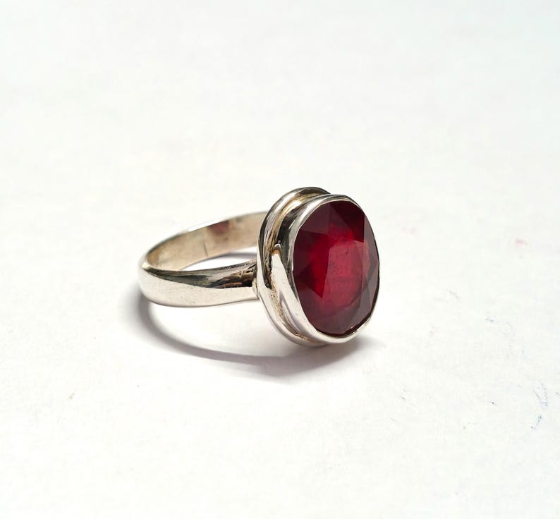 May include: A silver ring with a large, oval-shaped, deep red gemstone. The gemstone is set in a bezel setting, and the band is polished silver. The ring is shown on a white background.