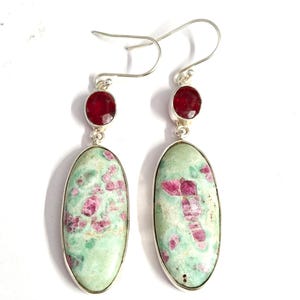 Pu&ograve; includere: Un paio di orecchini pendenti con pietre ovali verde chiaro con inclusioni rosa, incastonate in argento. Ogni orecchino ha una piccola pietra rossa rotonda nella parte superiore. Gli orecchini pendono da ganci d'argento.