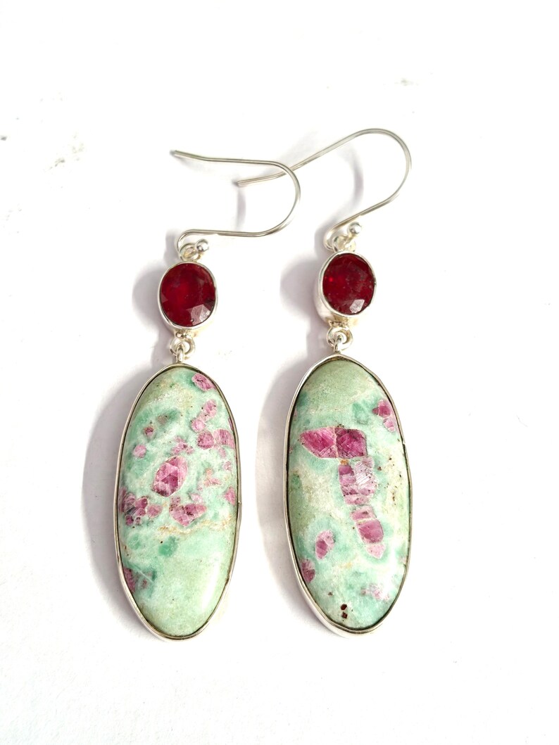 Pu&ograve; includere: Un paio di orecchini pendenti con pietre ovali verde chiaro con inclusioni rosa, incastonate in argento. Ogni orecchino ha una piccola pietra rossa rotonda nella parte superiore. Gli orecchini pendono da ganci d'argento.