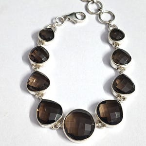 Könnte beinhalten: Ein silbernes Armband mit facettierten Rauchquarz-Edelsteinen. Das Armband hat dunkelbraune, unregelmäßig geformte Steine, die in einzelnen Silberfassungen gefasst sind und durch Silberglieder und einen Verschluss verbunden sind.