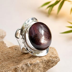 May include: A sterling silver ring featuring a large, oval-shaped, deep burgundy cabochon gemstone with a visible star-like chatoyancy effect. The metal band has a polished, hammered texture and a wide, layered setting that secures the stone.