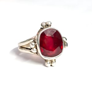 Peut inclure: Une bague en argent ornée d'une grosse pierre précieuse rouge foncé de taille ovale. La bague a un anneau fendu et des accents décoratifs en argent. La pierre précieuse est le point central, mettant en valeur sa couleur riche.