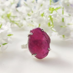 May include: A silver ring featuring a large, faceted, pink ruby gemstone. The ruby is set in a claw setting and has a natural, irregular shape.
