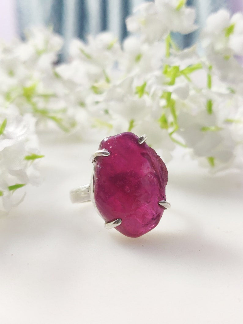 May include: A silver ring featuring a large, faceted, pink ruby gemstone. The ruby is set in a claw setting and has a natural, irregular shape.